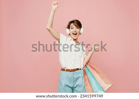 Young woman wear casual clothes hold shopping paper package bags listen to music in headphones raise up hand isolated on plain pastel pink color background studio. Black Friday sale buy day concept