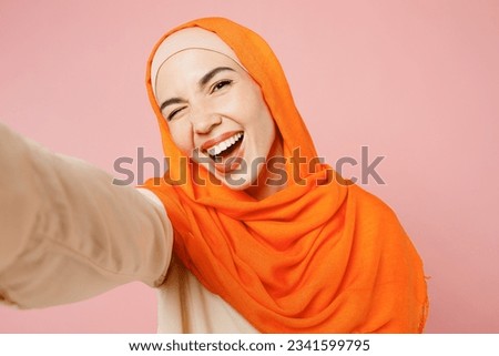 Close up young arabian asian muslim woman wear orange abaya hijab doing selfie shot pov on mobile cell phone wink isolated on plain pink background studio. Uae middle eastern islam religious concept