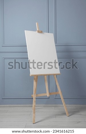 Wooden easel with blank canvas near grey wall indoors. Space for text