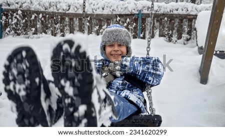 Cheerful laughing boy in warm hat and coat swinging on swing on playground covered with snow in winter.