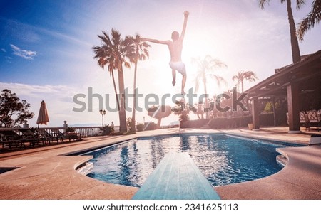 Boy jump from diving board to swimming pool, sunbeds and umbrellas lit with warm sunset light going through leaves of palm tree Royalty-Free Stock Photo #2341625113