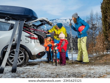 Family with father mother and two adorable little kids standing joking unloading ski, luggage by the open car trunk