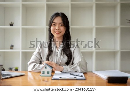 A beautiful Asian female real estate agent sitting at a table with a house model and contract on the table. property investment, apartment renting, house buying, residential