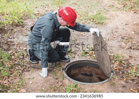 a man in overalls opened a sewer hatch and looks into a septic tank. Cleaning of sewers and drains. Royalty-Free Stock Photo #2341636985