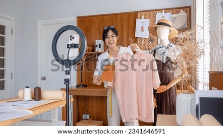 Young female designer or stylist takes center stage in her studio, live streaming an engaging styling session to online customers with smartphone showcases a shirt and color palettes.