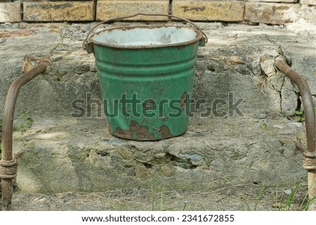 one rusty, iron green color old dirty retro empty bucket with a handle for water stands on concrete steps on the street in the afternoon in summer near a brick wall Royalty-Free Stock Photo #2341672855