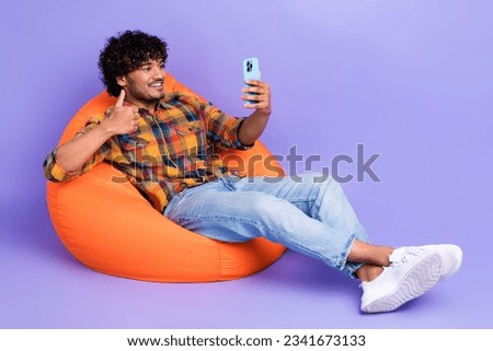 Full size photo of positive man sit on orange comfort bean bag hold smartphone showing thumb up isolated on purple color background