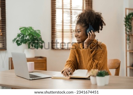 Smiling young african american woman in wireless headphones sitting at desk table working on laptop and writing letter in paper notebook, taking notes watching weninar Royalty-Free Stock Photo #2341674451