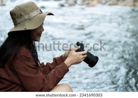 Side view of happy Asian woman with photo camera shooting tropical waterfall. Summer vacation Concept of wanderlust and adventure