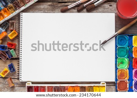Set of watercolor paints,  brushes for painting and blank white paper sheet of sketchbook on vintage wooden background. Top view.