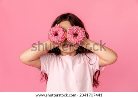 Portrait of a little smiling girl with curly hair and two appetizing donuts in her hands, closes her eyes with donuts, on a pink background, a place for text. Dieting concept and junk food. Royalty-Free Stock Photo #2341707441