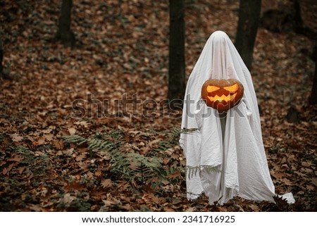 Happy Halloween! Spooky ghost holding glowing jack o lantern in moody dark autumn forest. Person dressed in white sheet as ghost with pumpkin standing in atmospheric evening woods. Boo!