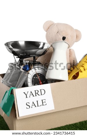 Sign Yard Sale written on box with different stuff on green grass against white background, closeup