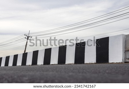 Close-up view of low angle concrete barriers painted in black and white painted on new paved road near electricity poles and trees and clouds in rural Thailand.