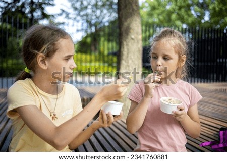 portrait of two cute blonde girls, they eat cold ice cream from paper cups. One tries a treat from another girl. A walk in the park, summer holidays and fun