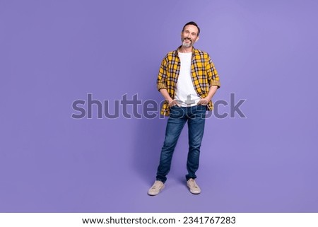 Full size photo of pleasant good mood man wear yellow plaid shirt jeans pants hold arms in pockets isolated on purple color background