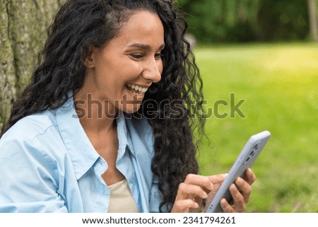 A cute Arab woman in the park holds a phone in her hands and writes a message, laughs and looks at the screen. Close-up portrait of a young girl in a blue shirt. Use device outdoors, online shopping
