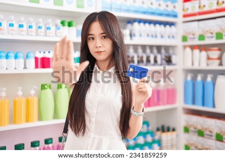 Young chinese woman shopping at pharmacy drugstore holding credit card with open hand doing stop sign with serious and confident expression, defense gesture 