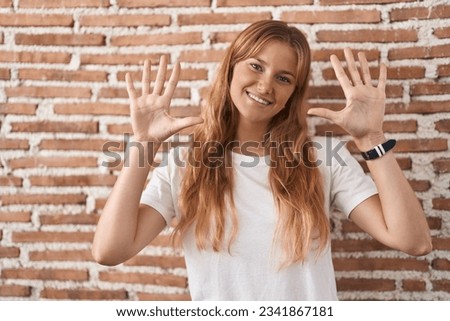 Young caucasian woman standing over bricks wall showing and pointing up with fingers number ten while smiling confident and happy. 
