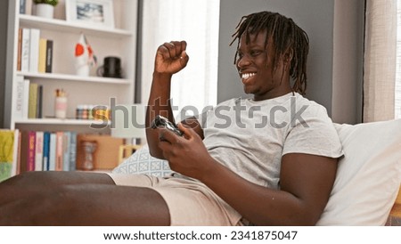 African american man playing video game sitting on bed celebrating at bedroom