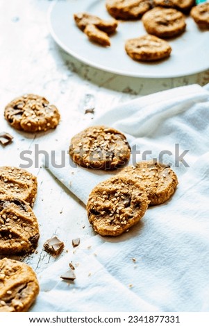 Overhead of gluten -free peanut butter garbanzo bean chocolate chip cookies with sesame seeds on white wooden surface with tray of cookies in the background.