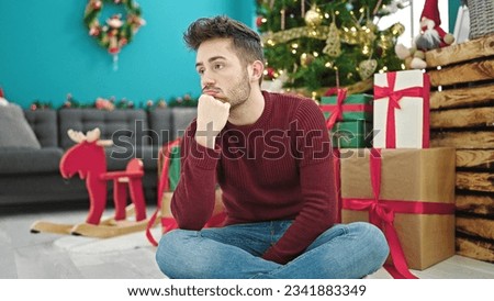 Young hispanic man celebrating christmas sitting on floor looking upset at home
