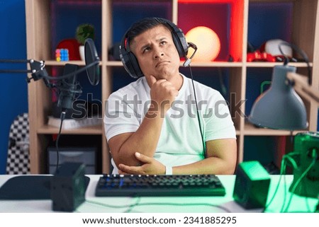 Young hispanic man playing video games with hand on chin thinking about question, pensive expression. smiling with thoughtful face. doubt concept. 