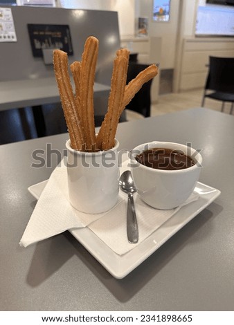 This is a photo of a plate of churros with a small cup of chocolate sauce. The churros are golden brown and freshly fried. The chocolate sauce is dark and thick. The background is a modern cafe.