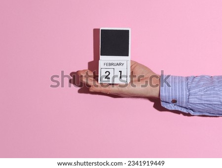 Man's hand in shirt holds wooden block calendar organizer with date February 21 on pink background with shadow