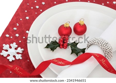 Christmas dinner place setting with white plate, red ribbon and baubles, holly and serviette with silver ring on table cloth.