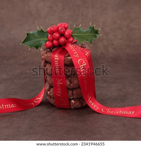 Chocolate chip cookie biscuit stack with holly and merry christmas red ribbon over brown lokta paper background.