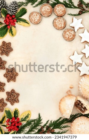 Christmas gingerbread biscuit selection and mince pie cakes forming a background border of holly and winter greenery over old parchment paper.