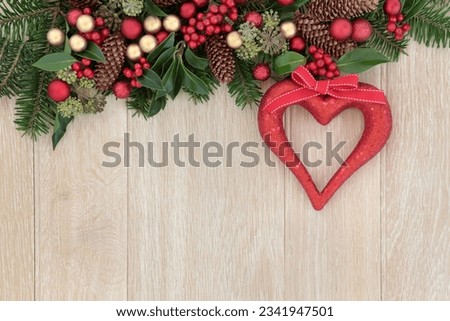 Christmas background border with red heart decoration, holly, baubles, mistletoe and winter greenery over light oak background.