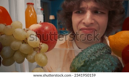 A man stands at the open refrigerator with healthy food. The upset man grimaces, not finding anything to eat. View from inside the refrigerator.