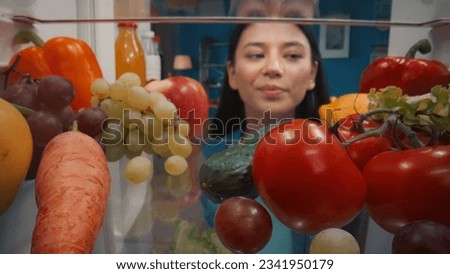 An Asian woman examines a refrigerator filled with healthy products: vegetables, fruits, berries. Satisfied woman standing by the fridge smiling. View from inside the refrigerator.