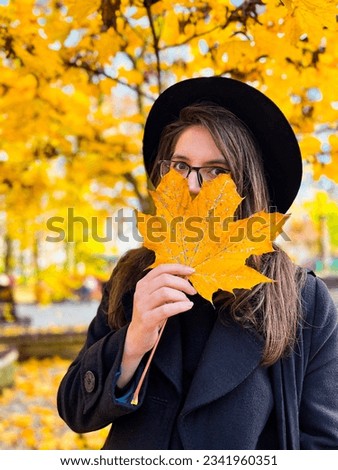 Wonderful brunette in a black hat, glasses, wearing black coat covers her face with a maple leaf in an autumn park. Many bright orange, yellow leaves around.