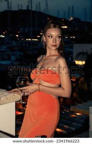A close-up portrait of a young woman in her early twenties wearing an orange dress, holding a glass filled with red wine