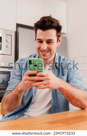 Vertical portrait of happy guy using his smart phone chatting sending messages on social media sitting at home kitchen and taking a break. Smiling young man playing a video game on cellphone. High Royalty-Free Stock Photo #2341973233
