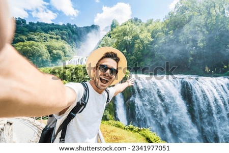 Handsome tourist visiting national park taking selfie picture in front of waterfall - Traveling life style concept with happy man wearing hat and sunglasses enjoying freedom in the nature 