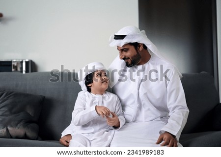 Arabs at home. Arabic father together with his son both wearing kandura Thobe Thawb while sitting on a coach. Emirati, Saudi, Oman, Qatar, Bahrain, Kuwait family concept Royalty-Free Stock Photo #2341989183