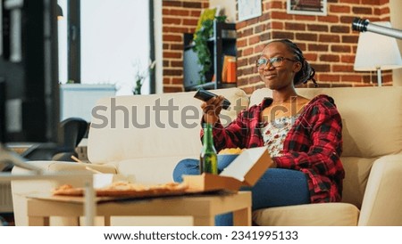 African american girl opening television to watch movie, eating chips aand drinking alcohol on couch. Smiling woman feeling happy with fast food meal in front of tv. Handheld shot.