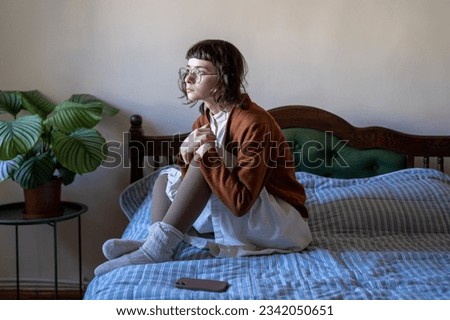 Thoughtful worried teen girl sits on bed sadly look at window waiting call, important message on smartphone. Lonely upset young woman holding knees in hand thinking about problem received bad news  Royalty-Free Stock Photo #2342050651