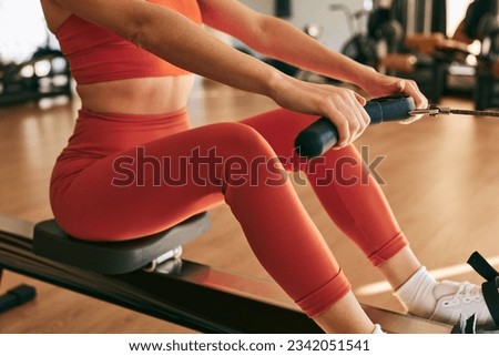 Crop anonymous female athlete in tight orange sportswear doing exercise with resistance band on rowing machine during workout in gym Royalty-Free Stock Photo #2342051541