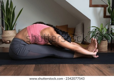 Young woman doing yoga stretching exercise, forward bend, paschimottanasana pose on mat at home. Royalty-Free Stock Photo #2342077543