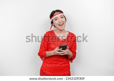 A portrait of a smiling Asian woman wearing red kebaya and headband and holding her phone, isolated by white background. Indonesia's independence day concept