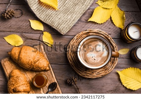 Autumn aesthetic concept. Cup of cappuccino coffee with croissants, candles, plaid and yellow autumn leaves on wooden background close up. Royalty-Free Stock Photo #2342117275