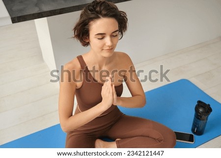 Young healthy beautiful woman in sports top and leggings practicing yoga at home, sitting in lotus position on yoga mat, meditating, smiling, relaxed, eyes closed, meditation concept.