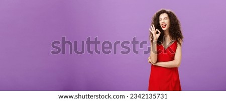 Cool enthusiastic caucasian female with curly hairstyle winking joyfully sticking out tongue and showing okay sign in approval and confirmation posing satisfied and confident over purple wall. Copy