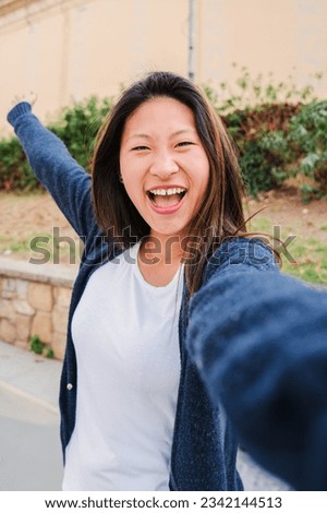 Vertical portrait of young asian brunette woman taking a selfie smiling and spreading arms. Teenage happy girl having fun shooting a individual photo looking at camera. Front view of positive female