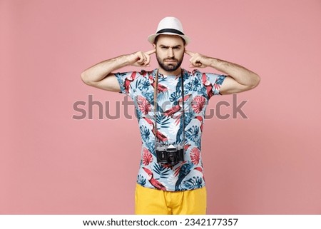 Concerned young traveler tourist man in summer clothes hat with photo camera covering ears with fingers isolated on pink background. Passenger traveling abroad on weekends. Air flight journey concept